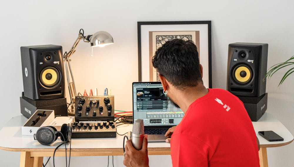 Man holding a microphone while using SoundID VoiceAI plugin in Pro Tools on a laptop, surrounded by studio monitors, audio gear, and headphones in a home studio setup