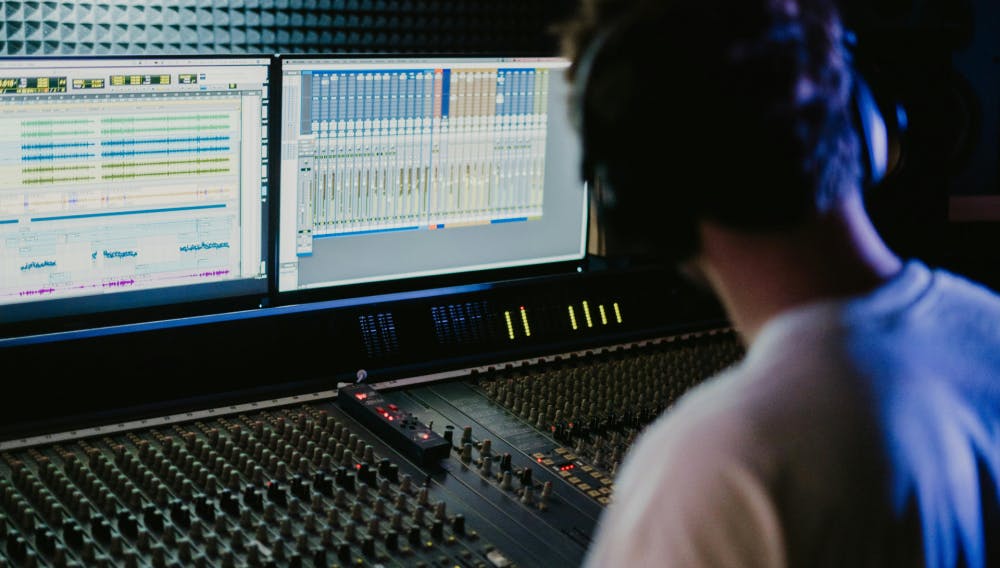 Audio engineer wearing headphones working at a professional mixing console with dual monitors displaying a digital audio workstation (DAW) interface