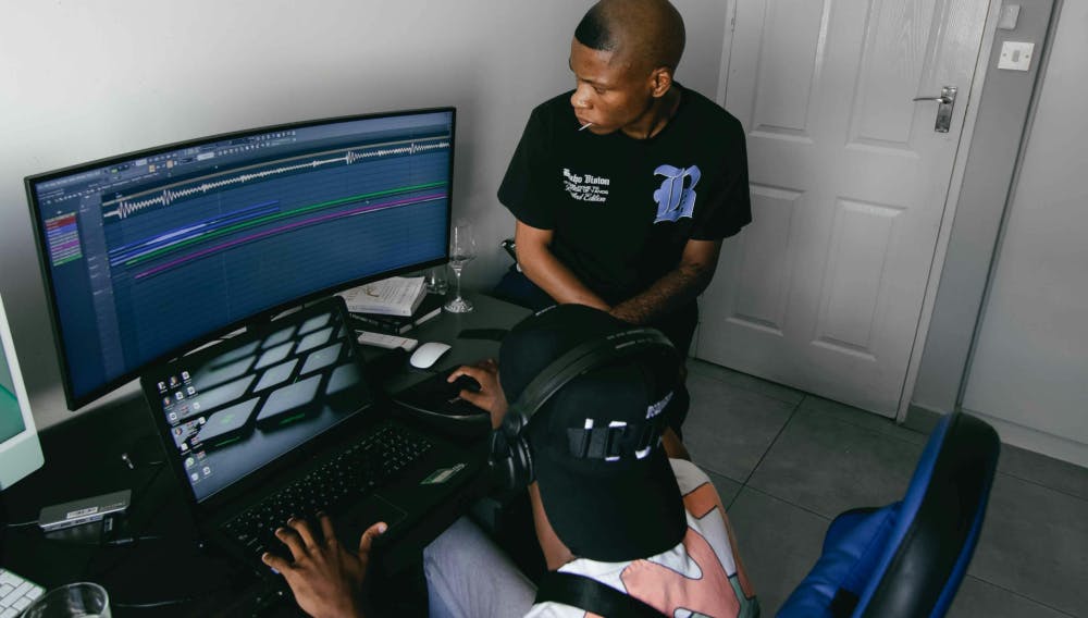 Two young music producers working on a beat in a home studio setup, with FL Studio open on a curved monitor and a laptop nearby.