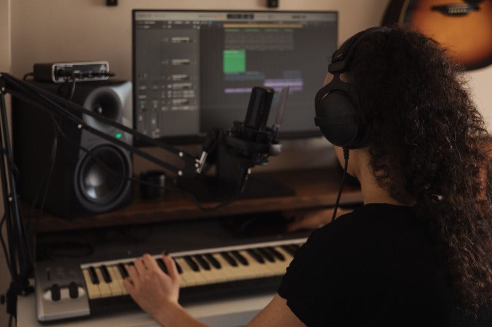 A person wearing headphones sits at a music workstation, playing a MIDI keyboard. A studio microphone is mounted on an adjustable boom arm, positioned in front of a computer monitor showing a digital audio workstation (DAW) interface. Studio monitor speakers and a wall-mounted guitar are visible in the background.