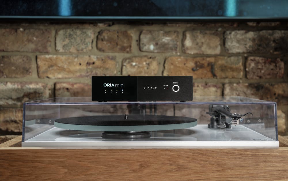 ORIA Mini hardware room-correction DSP sitting on top of a turntable dust cover in a brick-walled studio setting