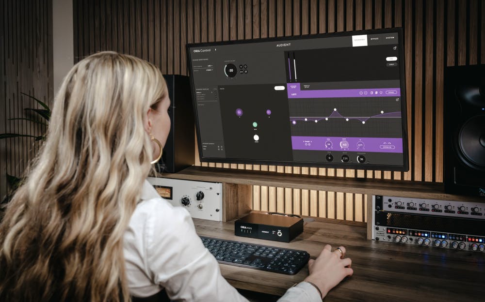 Female producer using ORIA Mini and ORIA Control software in professional studio with wooden acoustic treatment, monitoring setup, and audio interface equipment