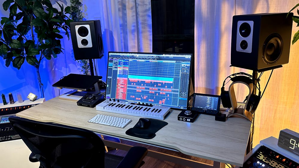 A close-up of a modern recording studio desk setup featuring a computer monitor running music production software, a MIDI keyboard, a studio monitor speaker on each side, a computer keyboard, a control interface, programable clock, and decorative crystals, surrounded by plants.