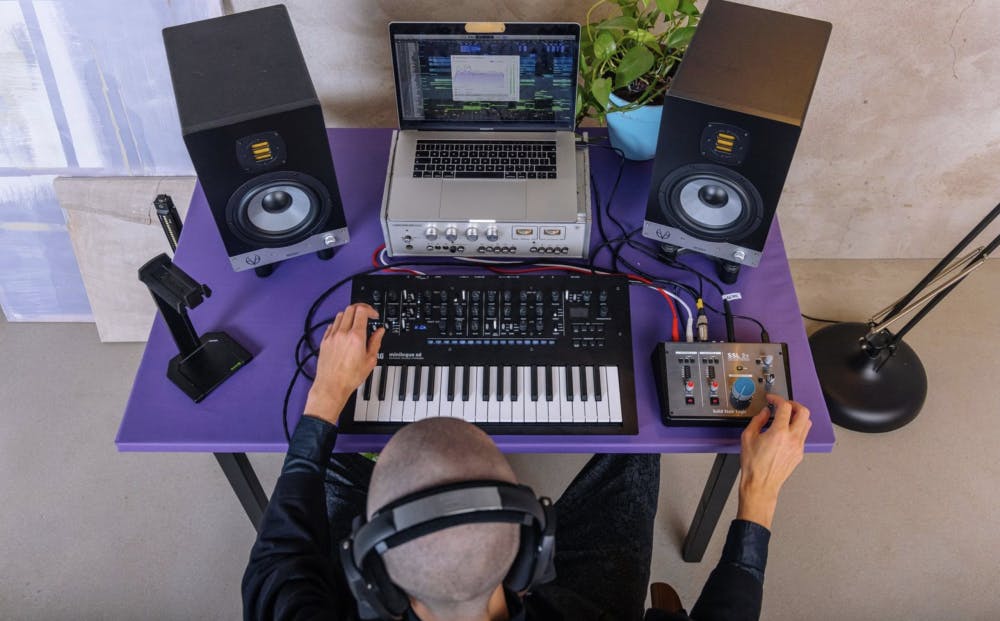 Overhead view of music producer working at home studio desk with stereo monitor speakers, synthesizer, audio interface, and laptop running DAW for binaural audio monitoring and music production
