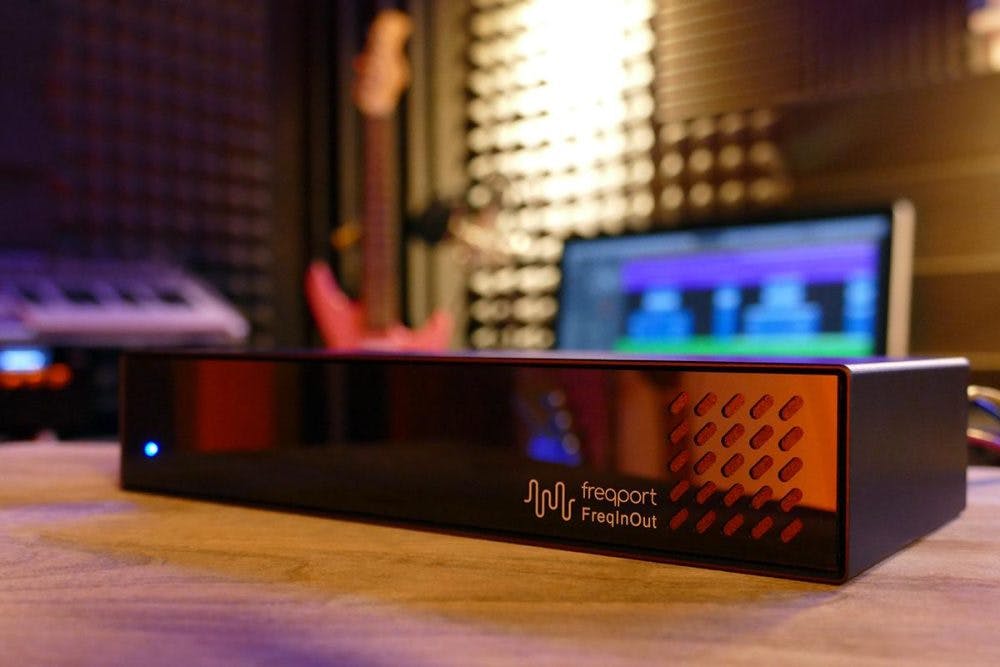 Freqport FreqInOut audio interface on a studio desk with acoustic treatment, electric guitar, MIDI keyboard, and DAW software visible in the background of a home production setup