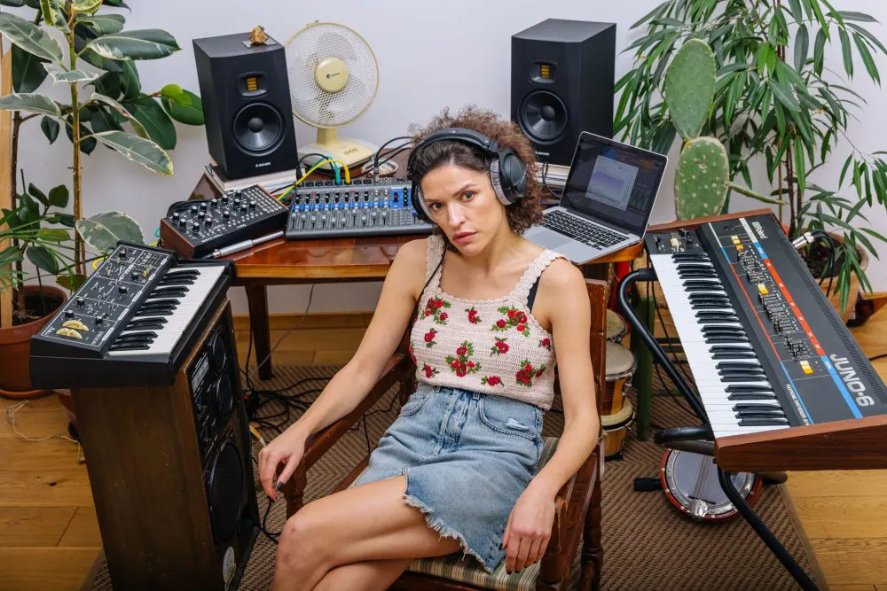 Female music producer wearing headphones seated in a home studio surrounded by synthesizers, studio monitors, and a mixing console