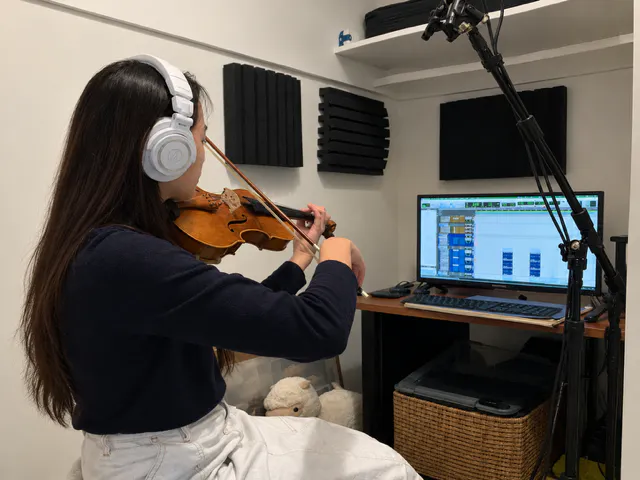 Violinist Myra Choo wearing headphones, recording into a microphone on a stand in a small home studio with acoustic foam panels and a DAW session on the monitor